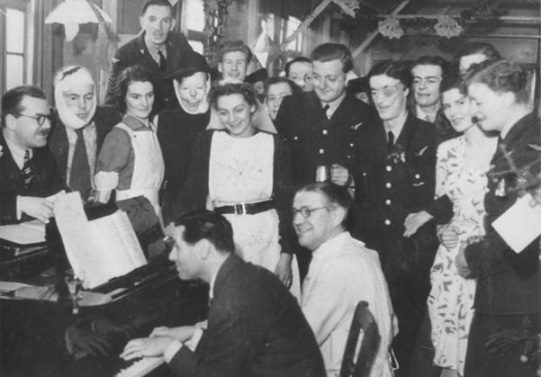 Smiling people (some wounded and bandaged) around a man at a piano