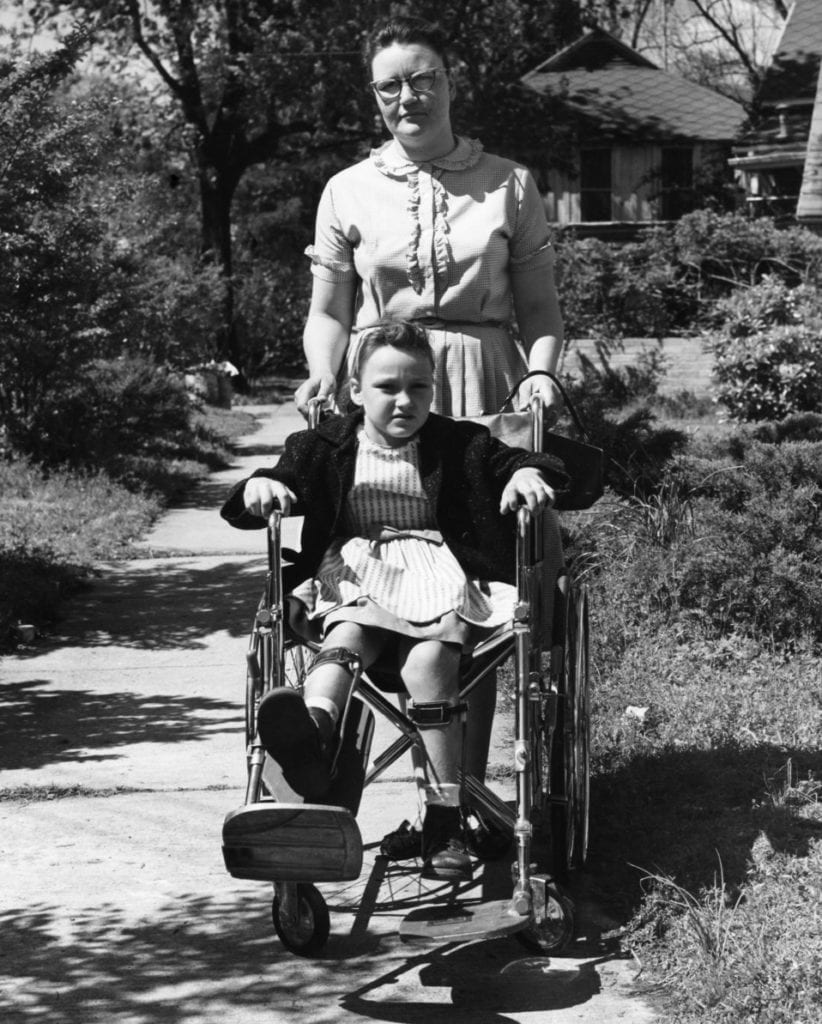 A young girl suffering from polio, seated in a wheelchair pushed by a woman