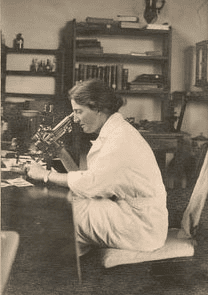 Black and white photo of Lucy Wills, who treated anemia with yeast extract, or marmite. Depicts a white woman with dark hair looking into a microscope.
