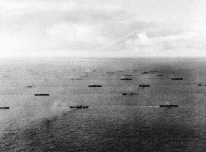 Black and white aerial photo of more than 30 ships crossing ocean.
